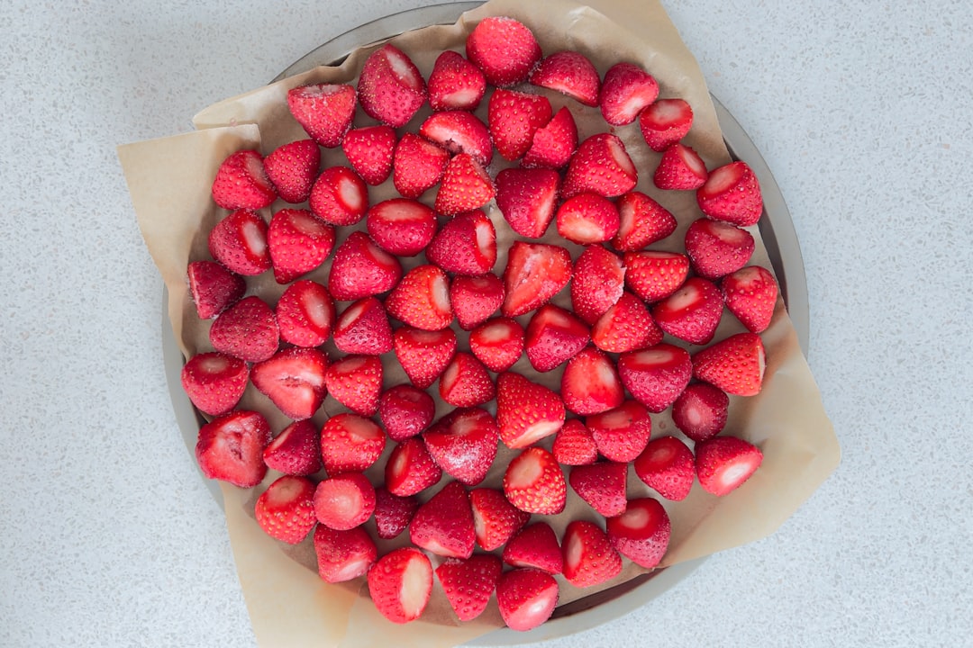 A high-angle, close-up shot captures a metal tray filled with halved, frozen strawberries. The vibrant red fruit is artfully arranged on parchment paper, creating a visually appealing and slightly frosty texture. The images minimalist composition and cool color palette lend themselves to various culinary and lifestyle applications.